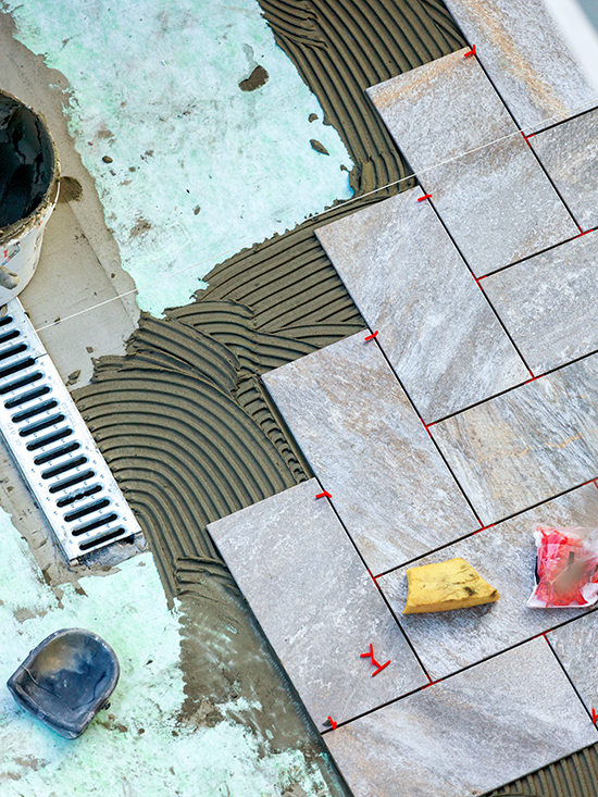 Close-up of stone tiles being laid with spacers on a mortar bed beside a drainage grate.