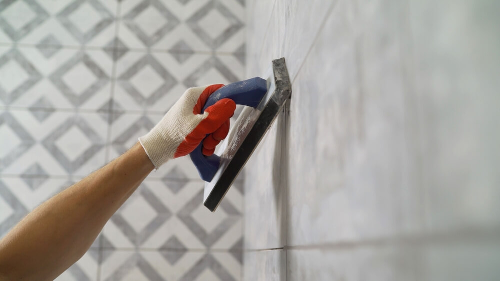 Hand with glove smoothing grout on tiled wall using a grout float tool.